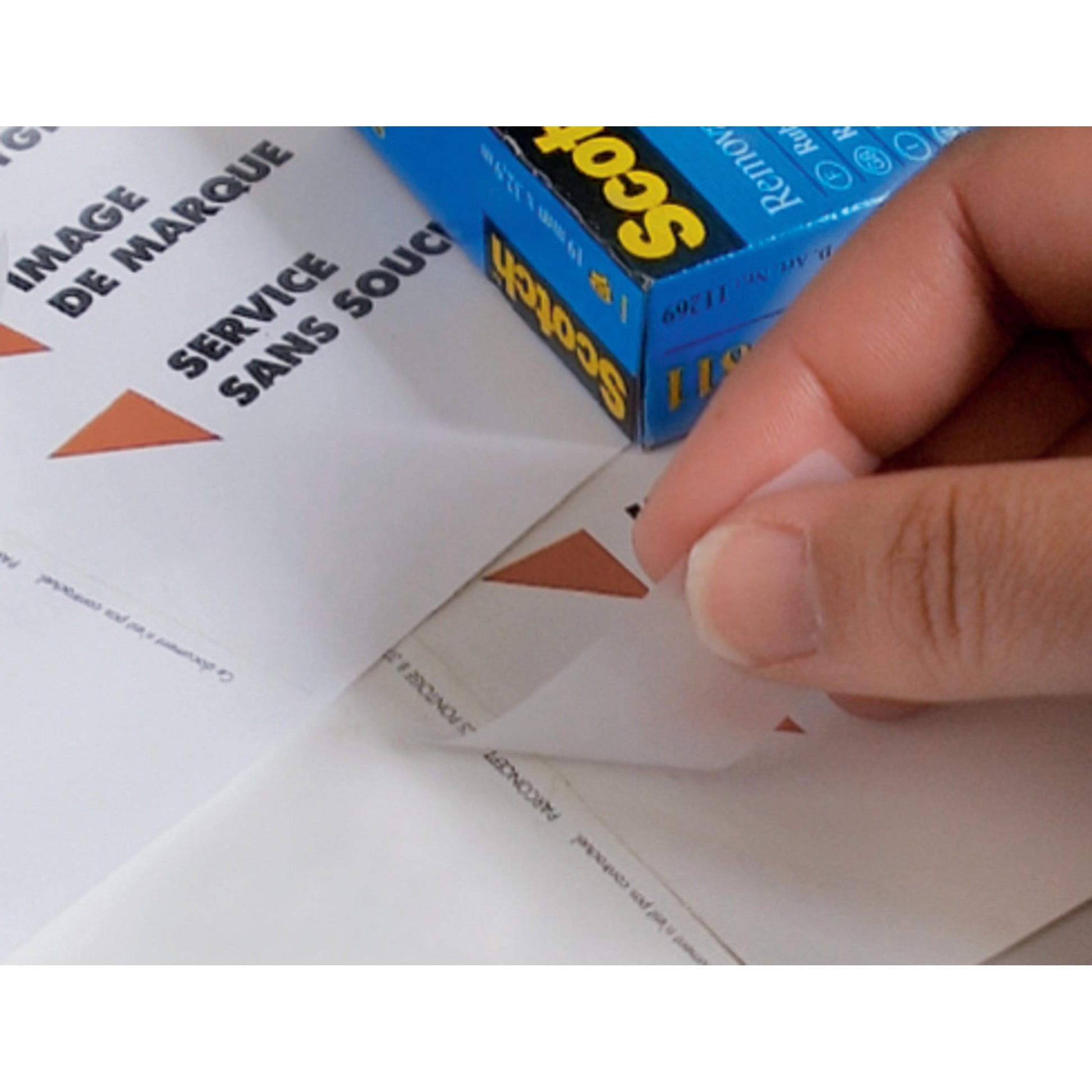 Eine Hand löst einen weißen Aufkleber mit einem braunen Dreieck von einem Blatt neben einem blauen Scotch® unsichtbares Klebeband, ablösbar, 19 mm x 33 m Karton der 3M Deutschland GmbH. Französischer und englischer Text ist teilweise auf dem Papier sichtbar.