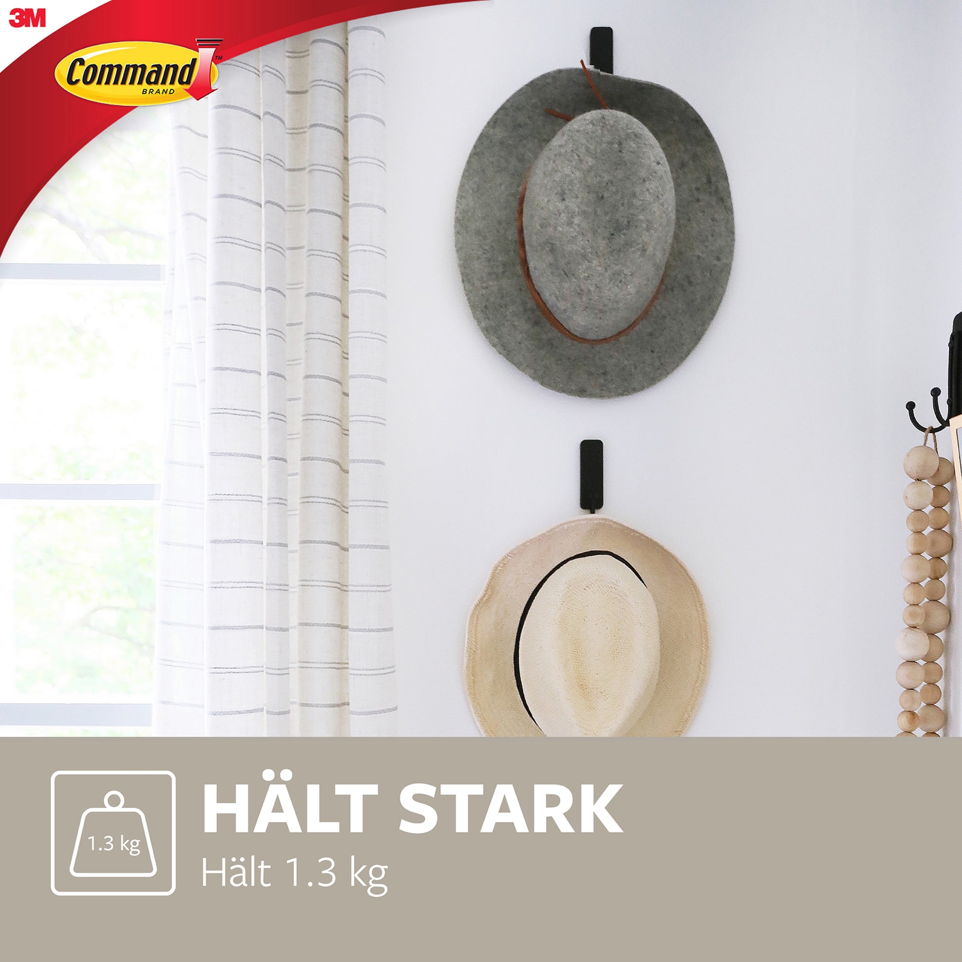 Zwei Hüte, einer grau und einer beige, hängen an einem 3M Mittelgroßer Command™ mattschwarzer Haken (17034B) neben einem Fenster mit weißen Vorhängen. Diese Wandhaken von 3M halten bis zu 1,3 kg und schützen Ihre Wände.