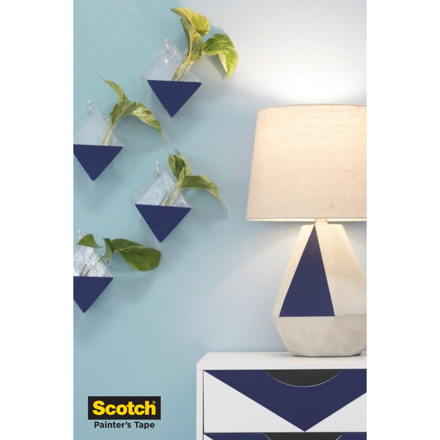 Eine blau-weiße, geometrische Kommode und eine Lampe stehen vor einer blauen Wand mit dreieckigen Pflanzgefäßen, alles geschützt mit Scotch® Malerabdeckband für empfindliche Oberflächen (36 mm x 41 m, 1 Rolle) von 3M Deutschland GmbH. Das Logo erscheint unten links.