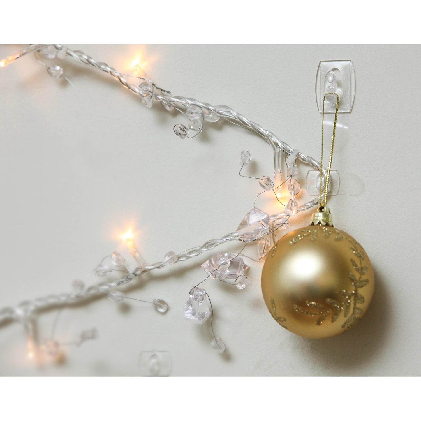 Ein goldenes Weihnachtsornament hängt an einem Command™ Mini-Haken, transparent, mit transparenten Strips, 17006CLR von 3M Deutschland GmbH, neben beleuchteten weißen Lichtern und klaren Kristallen an einer weißen Wand.