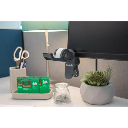 Pe birou: un dispenser de bandă adezivă 3M Scotch™ Clip & Twist cu bandă invizibilă Scotch® Magic™ (19 mm × 8,89 m), o lampă, o foarfecă într-un suport, două role suplimentare de bandă adezivă Scotch®, un pahar cu ace și o mică plantă suculentă în ghiveci.