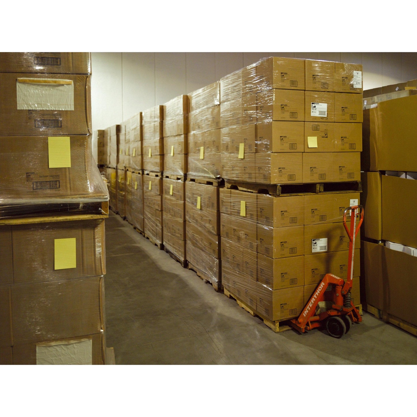 Într-un depozit sunt stive de notițe Post-it® Super Sticky în format mare, Ultra Galben (101 x 152 mm, 90 de foi/bloc, 6 blocuri), de la 3M Deutschland GmbH, ambalate pe paleți. Un transpalet roșu se află lângă ele.