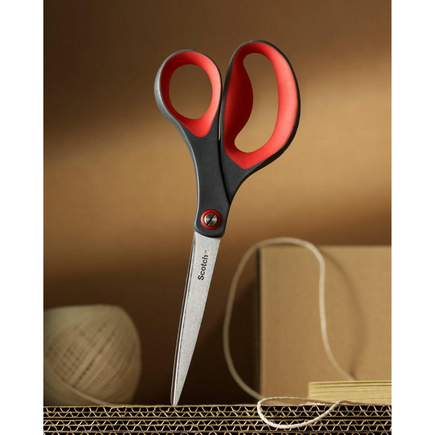 Ein Paar rote Scotch™ Präzisionsschere von 3M Deutschland GmbH ist aufrecht in Wellpappe geklebt, im Hintergrund sind beigefarbene Schnüre und ein Karton zu sehen.
