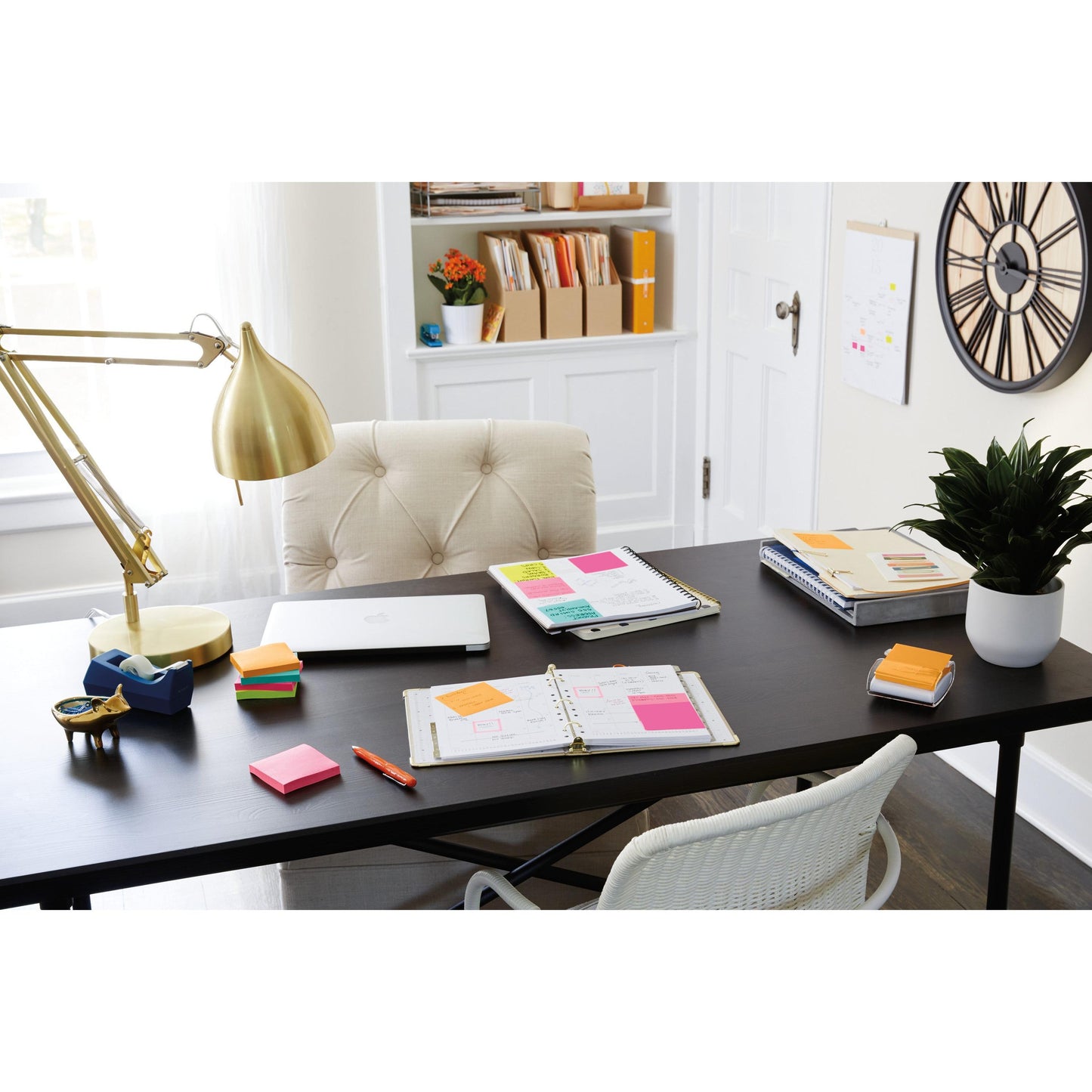 Pe un birou negru se află un laptop, pixuri, o lampă aurie, o plantă, articole de birou și Post-it® Super Sticky Notes 654-6SS-PPNK de la 3M Deutschland GmbH (roz, 76x76mm), lângă un scaun bej; cărți și cutii umplu rafturile albe din spate. În partea dreaptă este agățat un ceas de perete.