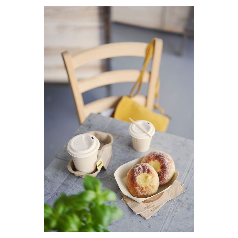 Pe o masă se află două produse de patiserie presărate cu zahăr pe un platou și două pahare de cafea Duni „Sweet Bagasse Imprimat” cu capac de la Duni GmbH. O geantă galbenă atârnă de un scaun de lemn, în timp ce o plantă verde parțial vizibilă în prim-plan subliniază subtil tema sustenabilității.